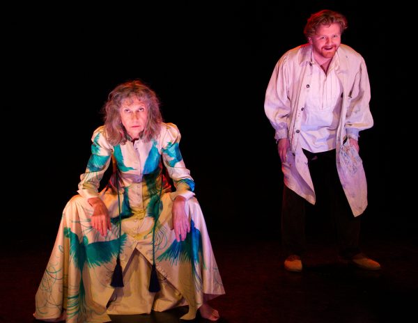 An eye-level medium shot, set against a stark black background, captures two actors in what appears to be a theatrical performance. To the left, an older woman with grayish, wavy hair sits facing the viewer in a slightly crouched position, her gaze directed upwards. She wears a long, cream-colored dress adorned with splashes of blue and green. Tassels accent the front of the dress. On the right stands a younger man, also gazing upwards with a smirk. He is clad in a painter’s smock over a white shirt and dark pants. The smock is visibly marked with paint stains. The lighting casts a vibrant red hue across the scene, creating a contrast with the figures and enhancing the dramatic atmosphere.