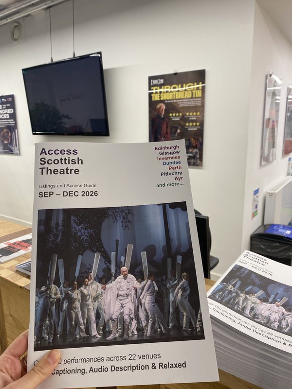 A hand holds a white “Access Scottish Theatre” listings guide in the foreground the reception of a theatre venue. The guide is labeled “SEP–DEC 2026” and lists Scottish locations including Edinburgh, Glasgow, Inverness, Dundee, Perth, Pitlochry, and Ayr. Its cover features a dramatic stage photograph of performers dressed in white, holding tall silver props around a central performer in a dark stage setting. Additional copies of the guide are stacked on a counter to the right. There is a TV screen and play poster in the background.